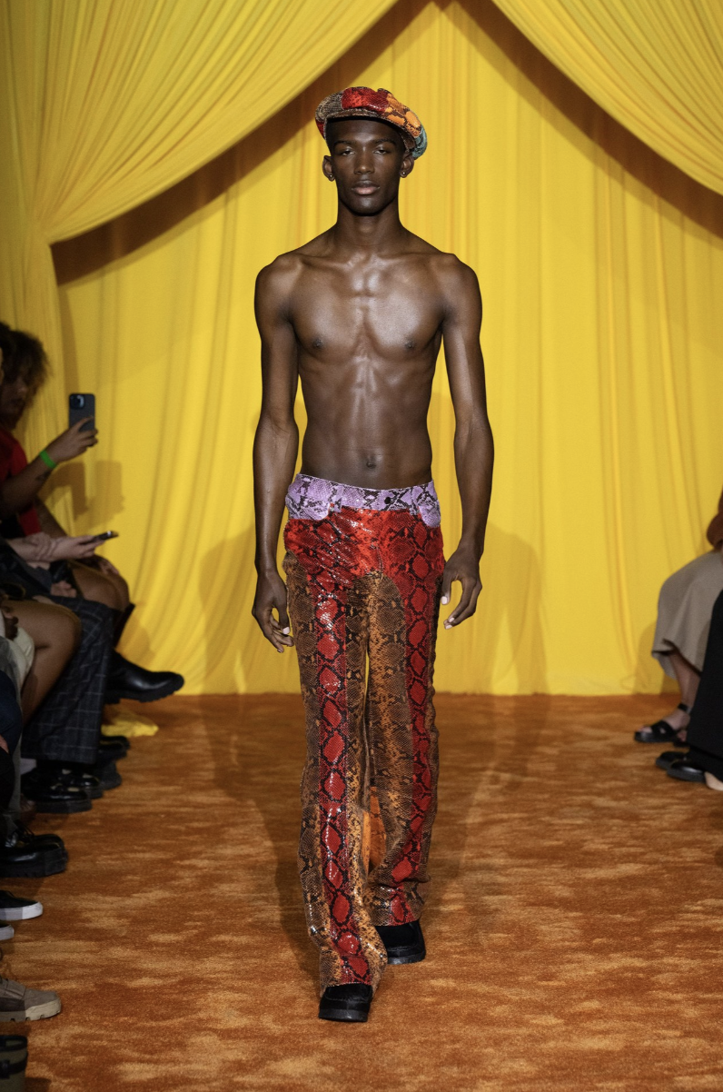 Slender male model with deep skin wearing only snakeskin pants and a snakeskin hat walking on a runway against a yellow backdrop